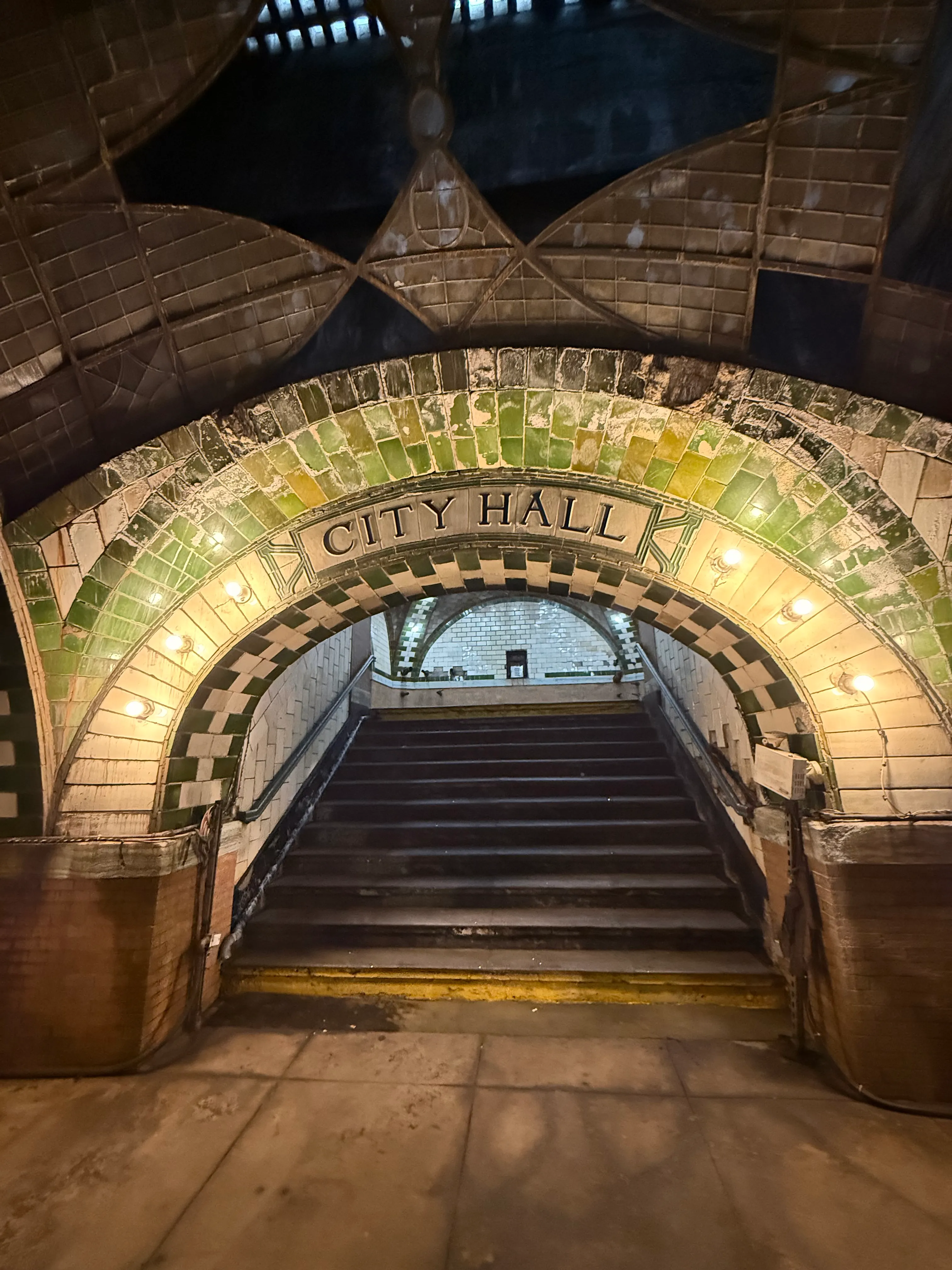 Beautiful, old station sign saying "City Hall" in front of the stairs going up and out of the station. The words are placed in a green-and-white tile arch with embedded lightbulbs. There&#x27;s a hint the main platform skylight visible as well.